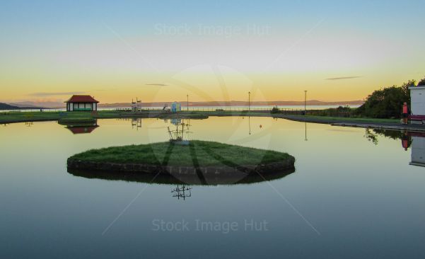 Little boating pond like a sheet of glass in early morning as sun begins to rise lighting up distant hills across the sea